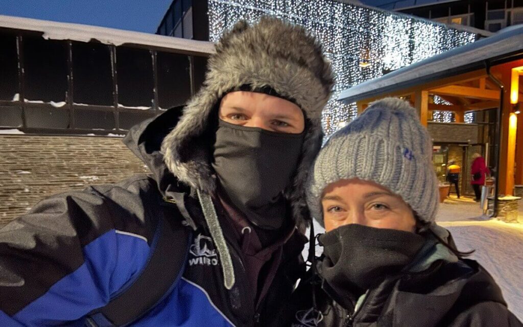 My wife and I in warm winter clothing, with my trapper hat and ski masks, in an outdoor evening setting, with sparkling lights in the background and snow on the ground.